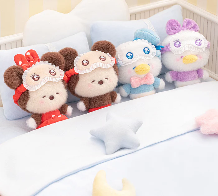 Four Disney squinting sleepwear blind box plushies in a crib with decorative pillows and a blanket. Featuring Mickey Mouse, Minnie Mouse, Donald Duck & Daisy