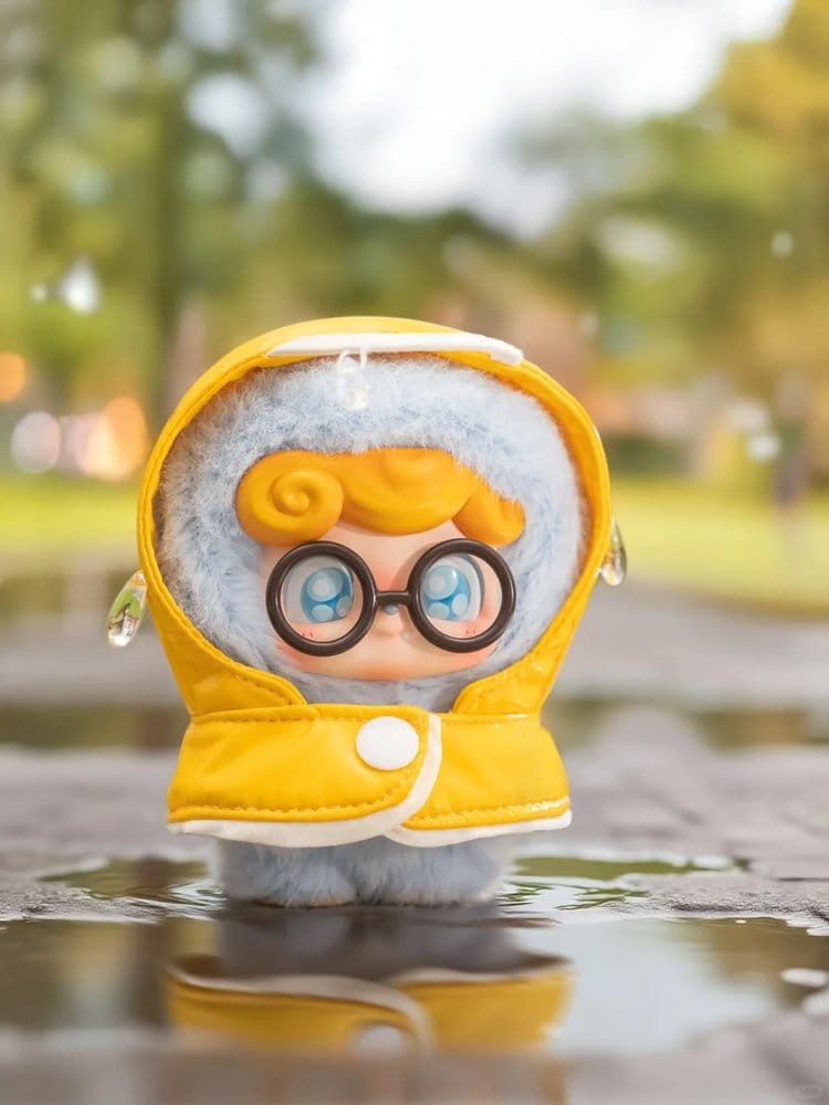 Small Q kid plush toy with a yellow raincoat and glasses standing on a reflective surface with a blurred natural background.