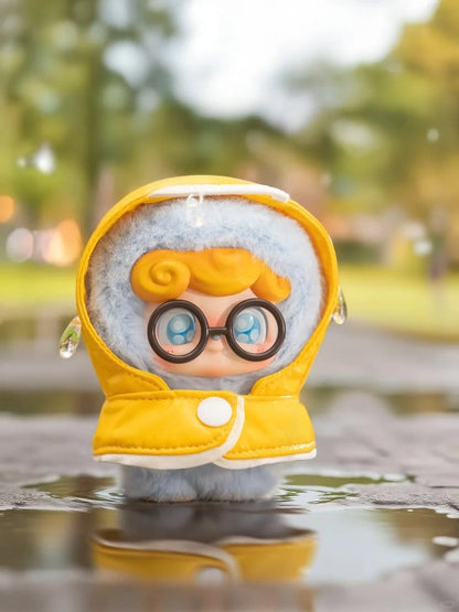 Small Q kid plush toy with a yellow raincoat and glasses standing on a reflective surface with a blurred natural background.